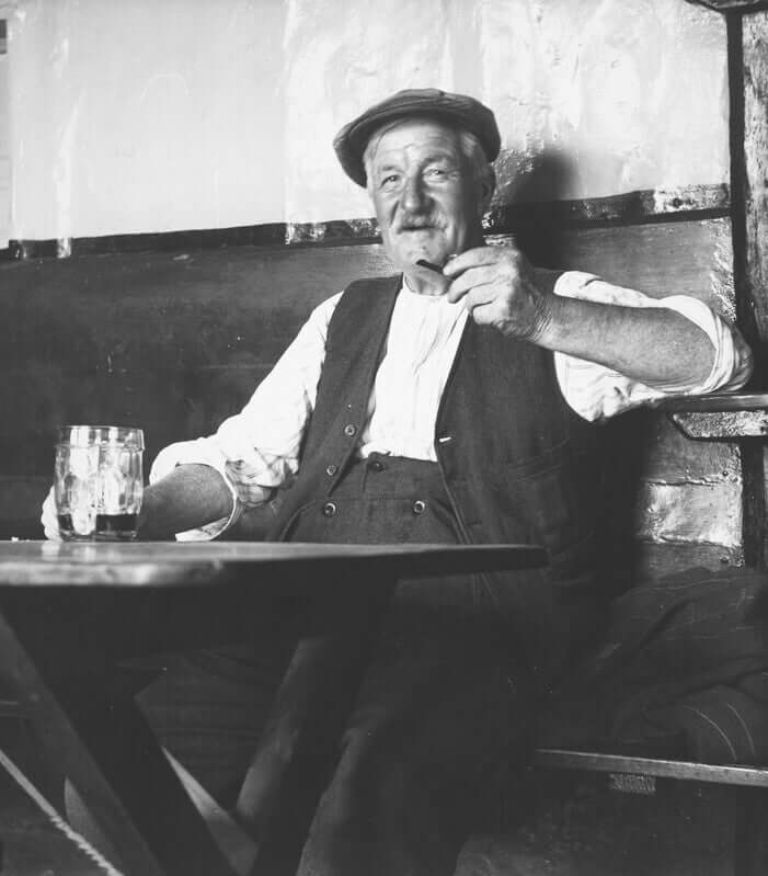 Elderly man relaxing at a table, enjoying a drink and smoking a pipe, vintage black and white photography.