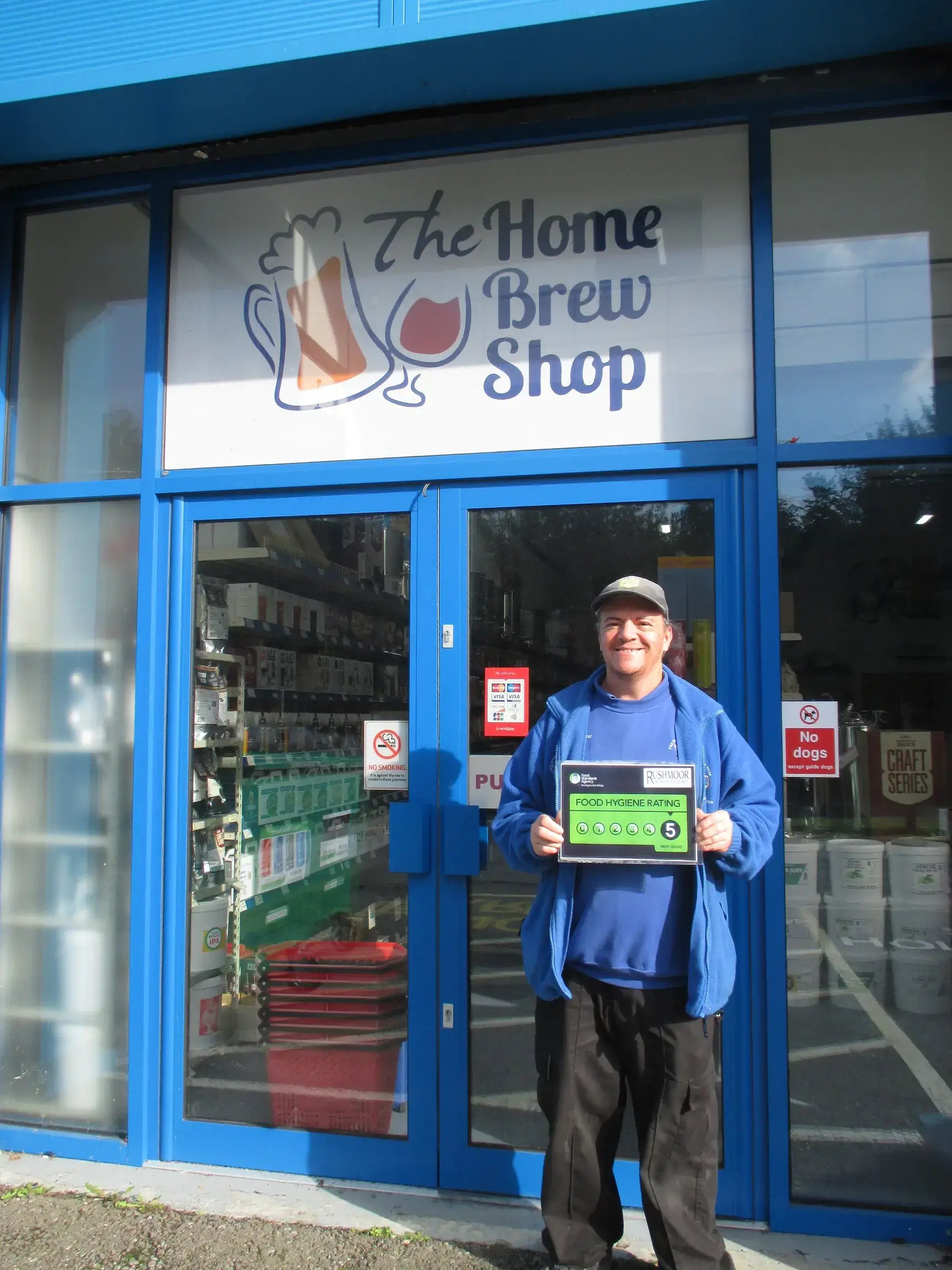 Smiling man holding a sign in front of The Home Brew Shop entrance with colorful signage.