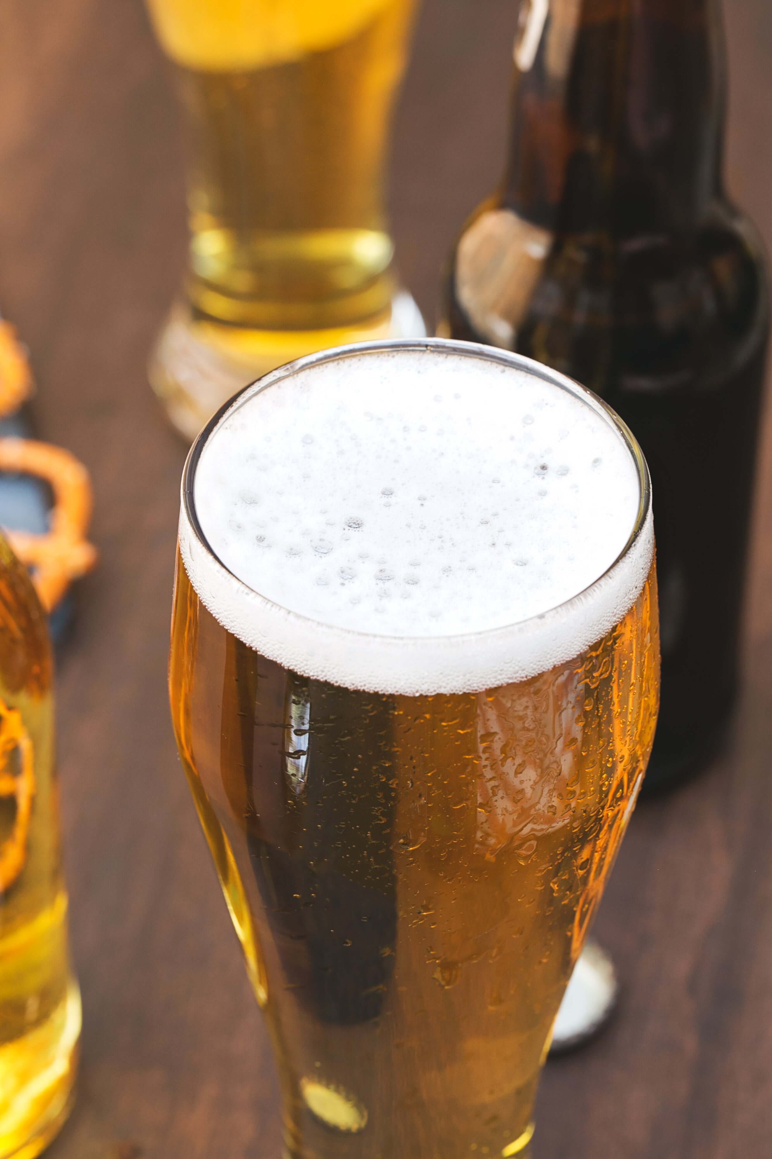 Close-up of a frothy beer in a glass, surrounded by bottles and other beer glasses on a wooden surface.