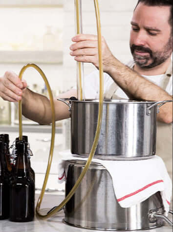 Man brewing beer, transferring liquid with a hose from a pot to bottles in a kitchen setting.