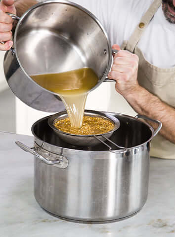 Chef pouring broth through a strainer into a stainless steel pot, showcasing the cooking process.